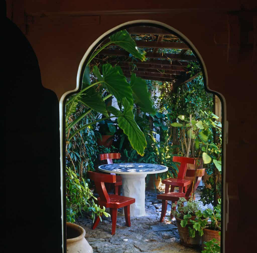 A round pedestal table with a mosaic tiled top is surrounded by a group of chairs designed by John Stefanidis under a pergola in the garden at his home in P