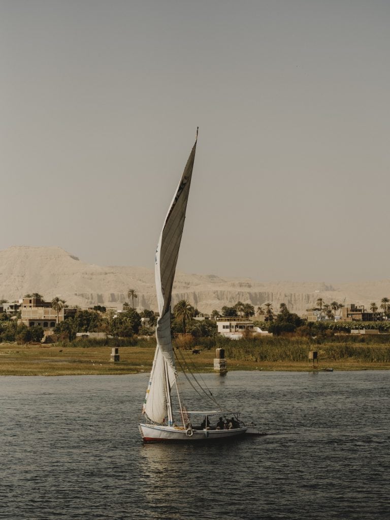 A felucca sailing boat on the Nile near Luxor, palm trees, mountains and the desert beyond.